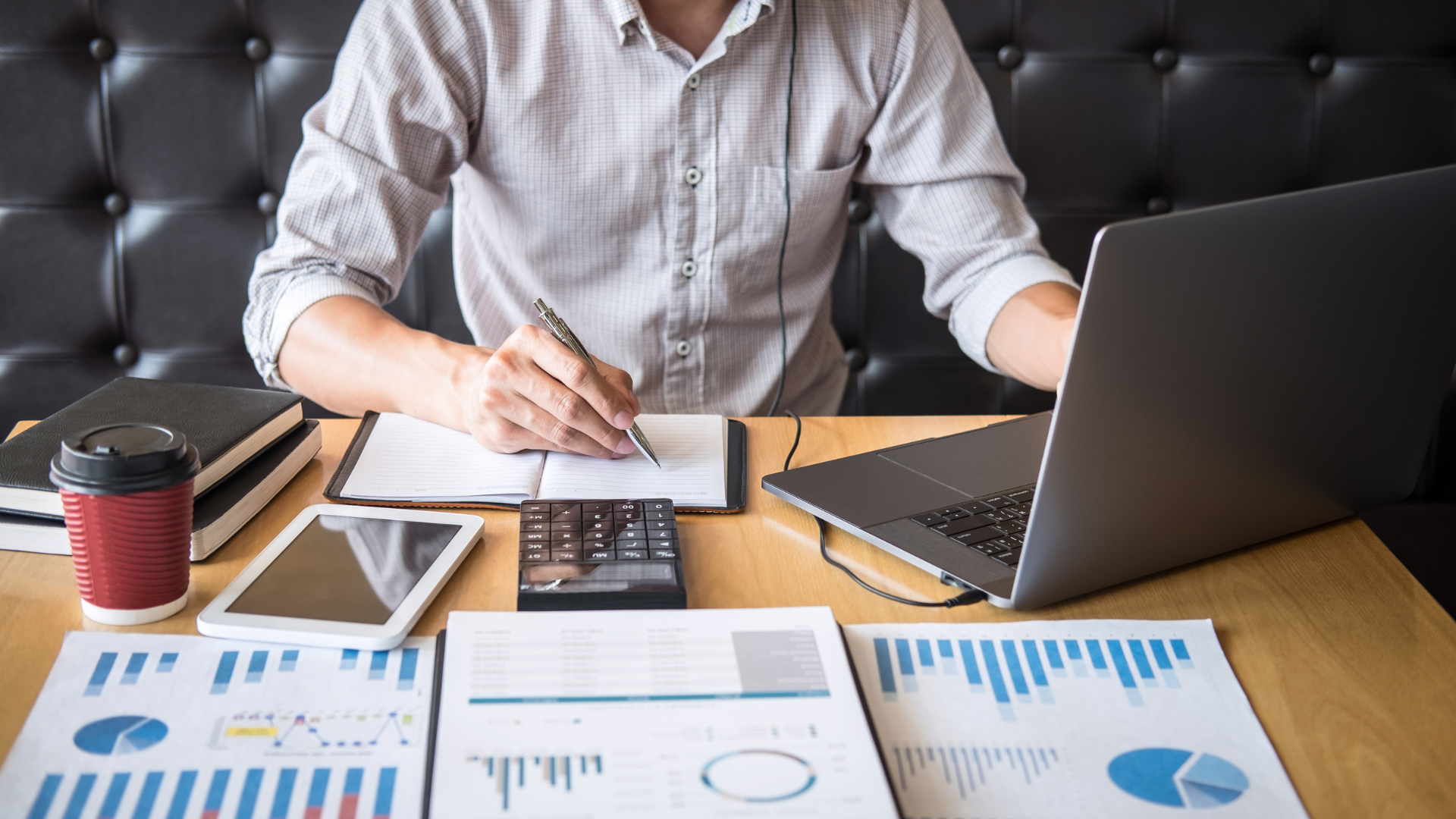a person sitting in front of a desk, looking at a laptop and financial charts
